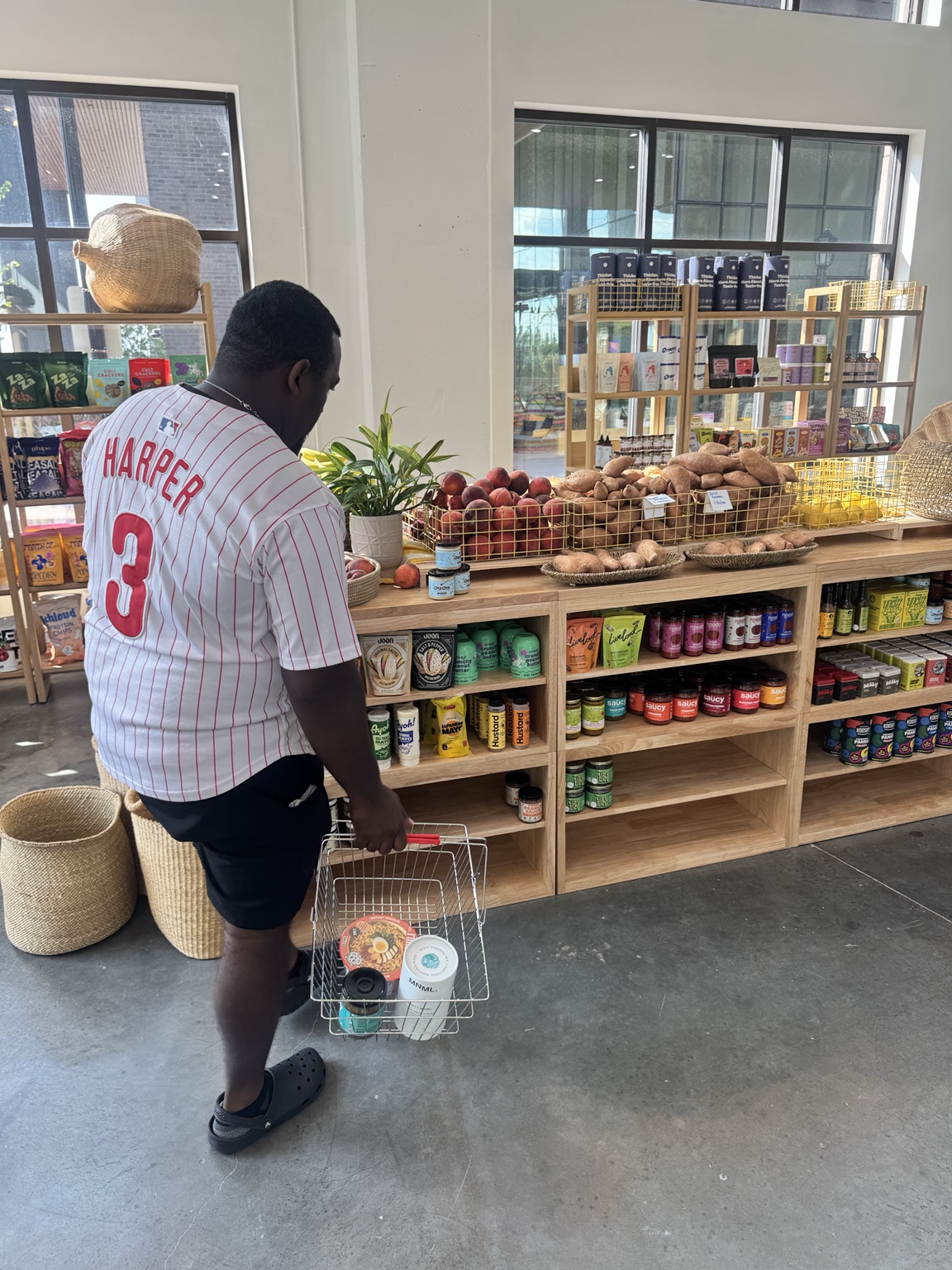 A shopper with a basket standing near colorful grocery shelves and fresh produce at Nelly's Grocery and Wine.